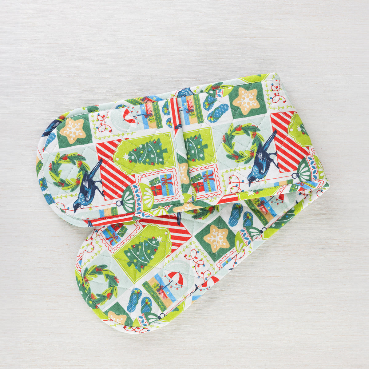 Christmas Wishes Kiwi Christmas double oven glove featuring a colourful patchwork-style print with Christmas trees, native birds, wreaths, ornaments and gifts.