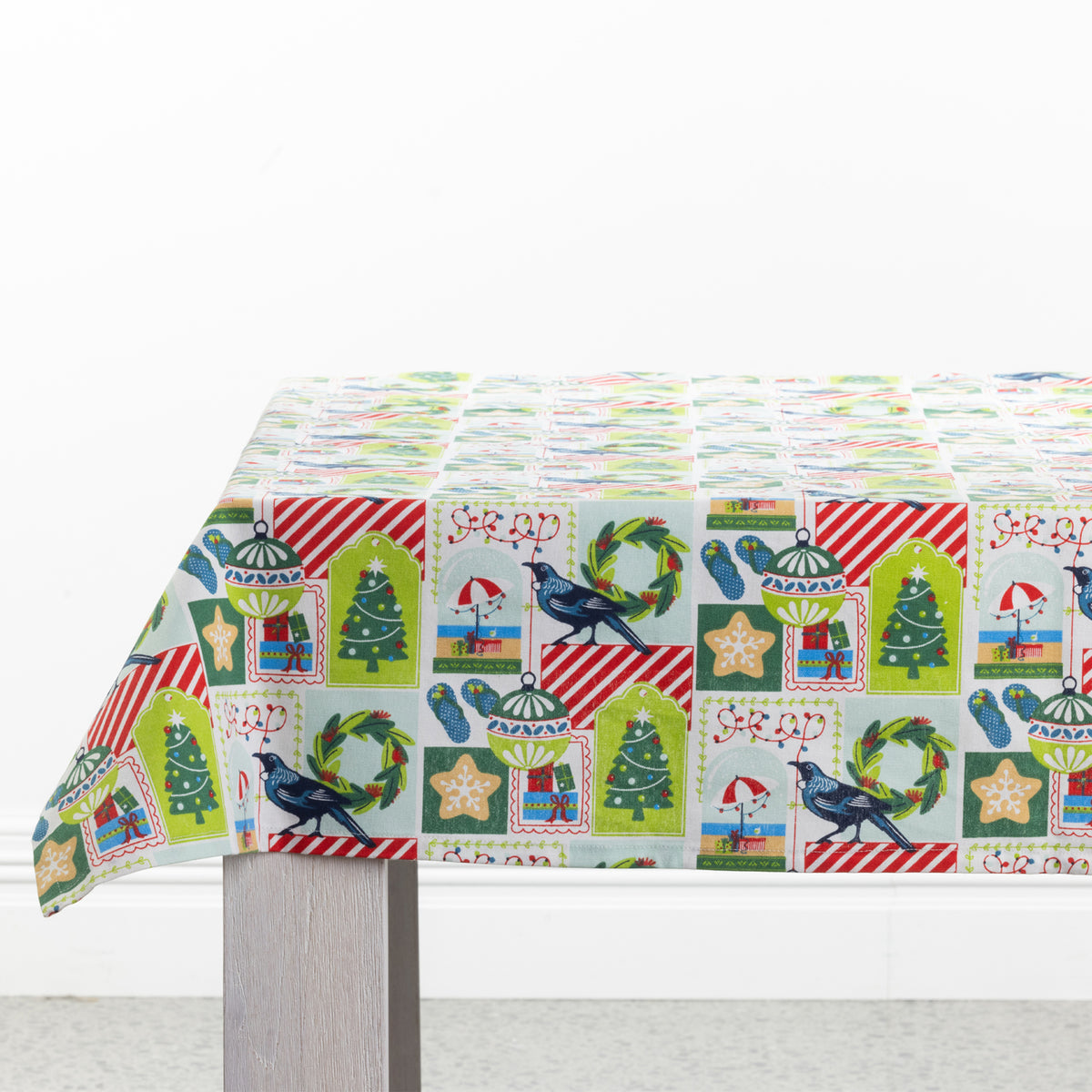 Christmas Wishes Kiwi Christmas Table Cloth draped over a table, featuring a colourful patchwork-style print with Christmas trees, native birds, wreaths, ornaments and festive details.