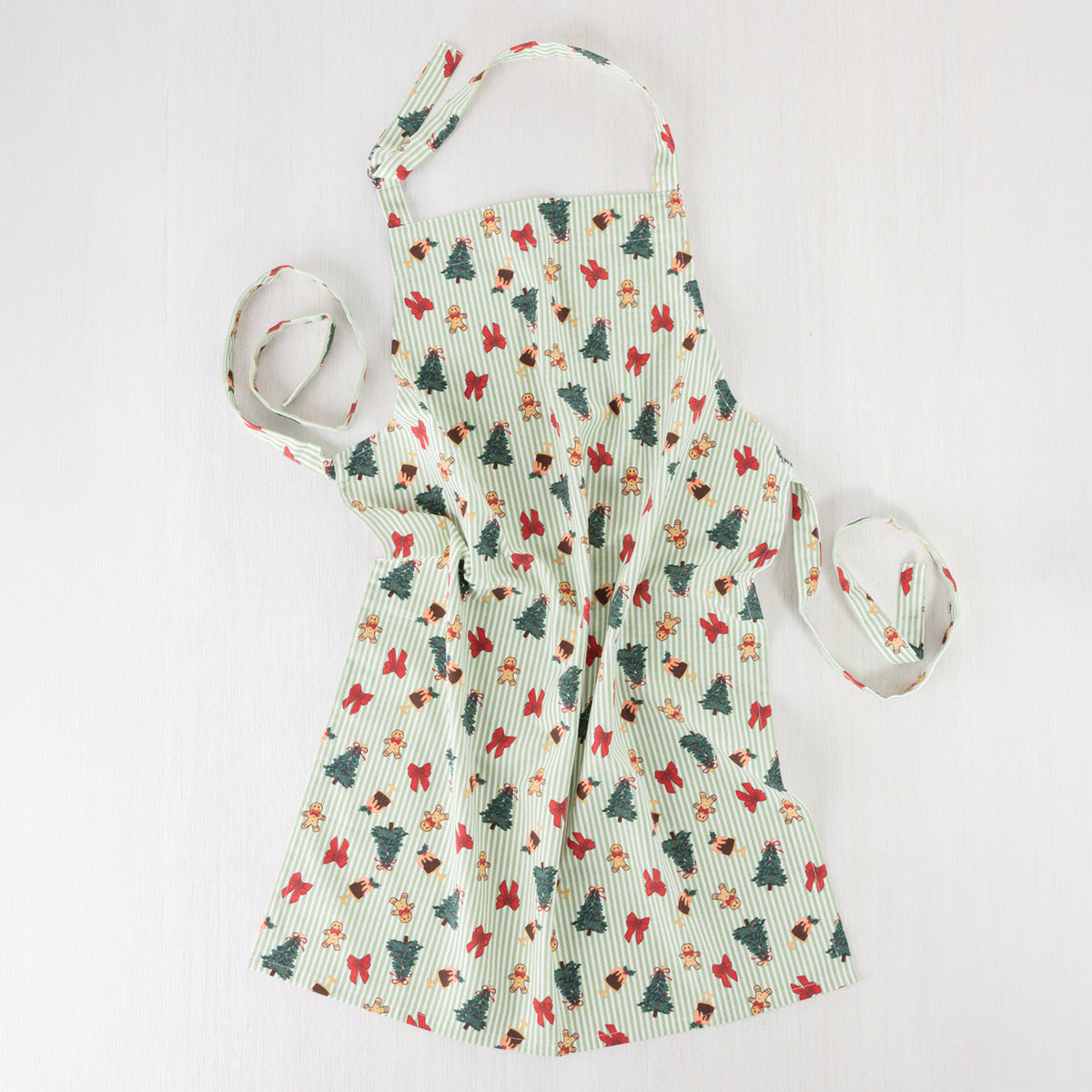 Christmas Wishes Classic Christmas Apron laid flat, featuring fine green and white stripes with festive Christmas trees, gingerbread people, red bows and Christmas puddings.