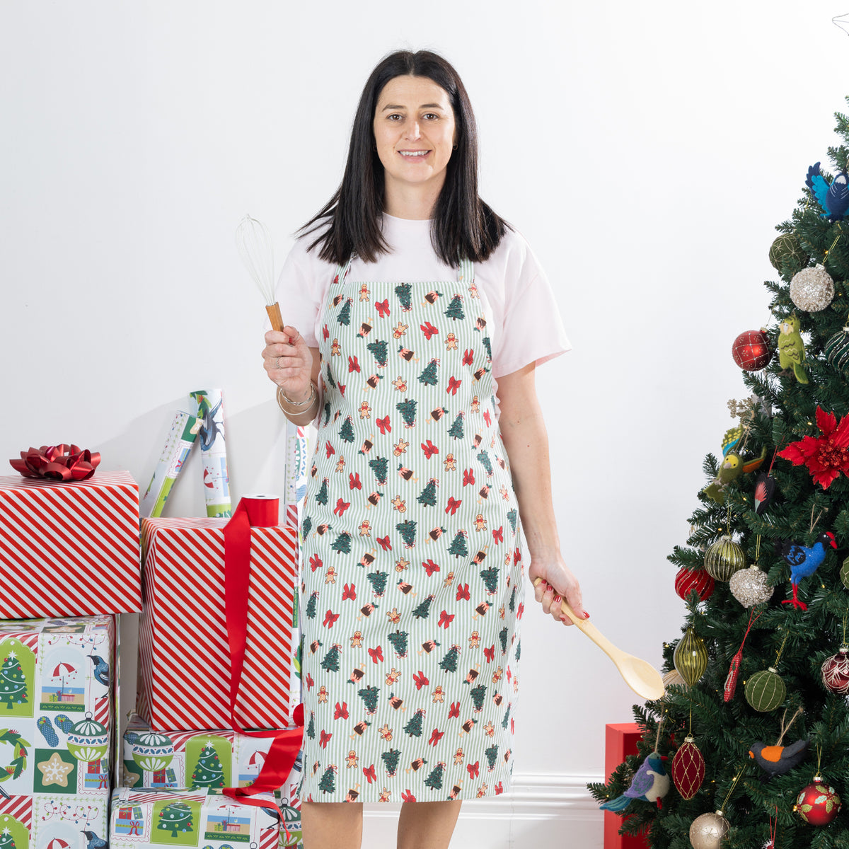 Christmas Wishes Classic Christmas Apron worn in a festive Christmas setting, showing the green striped print with Christmas trees, gingerbread people, bows and puddings.