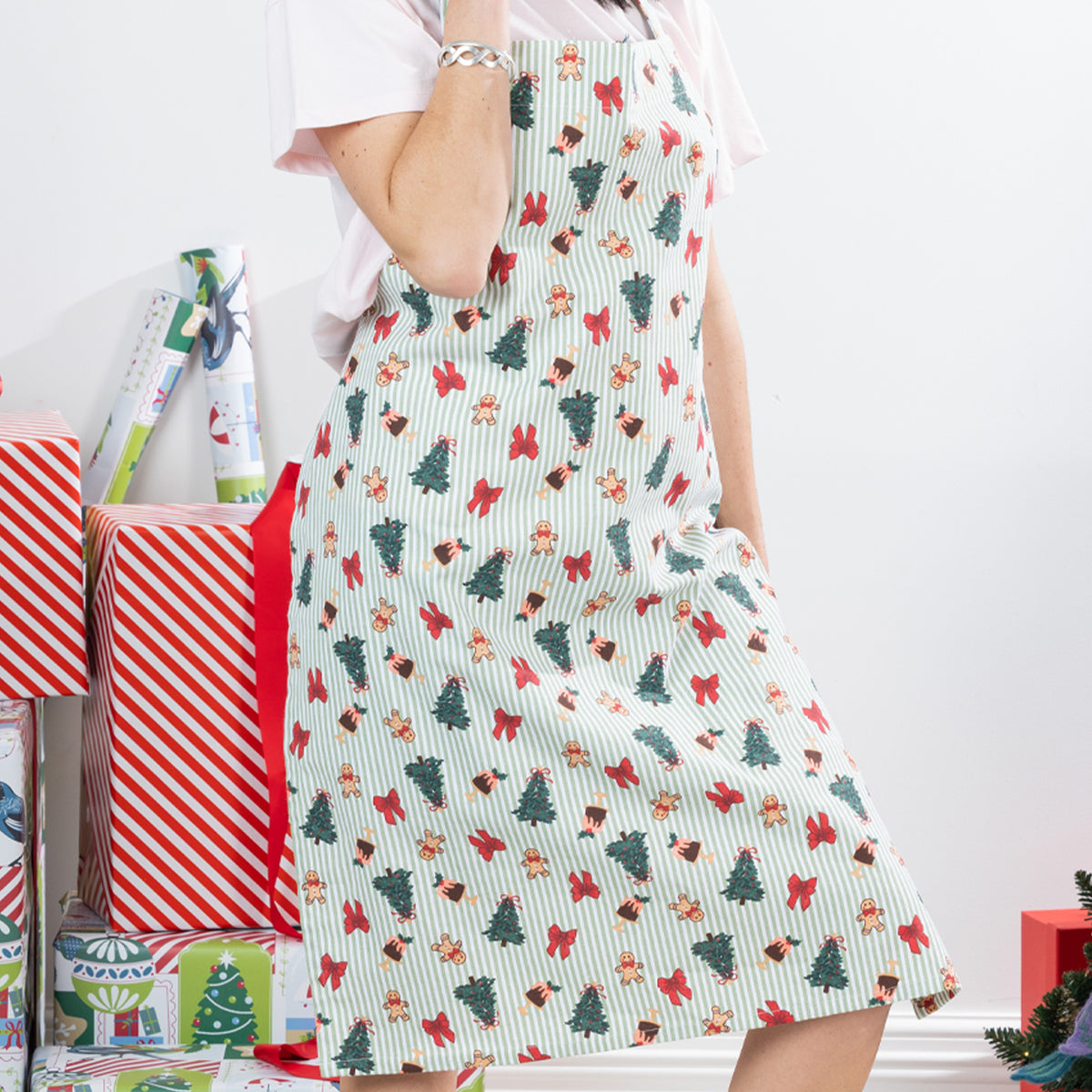Christmas Wishes Classic Christmas Apron worn in a festive Christmas setting, showing the green striped print with Christmas trees, gingerbread people, bows and puddings.