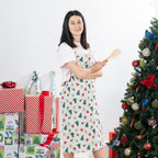 Christmas Wishes Classic Christmas Apron styled in a Christmas setting, worn with baking utensils beside a decorated tree and wrapped gifts.