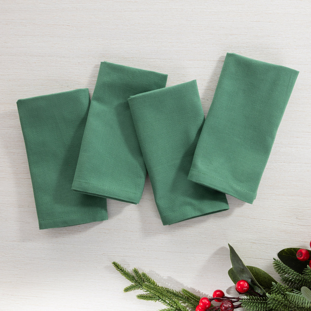 Christmas Wishes Classic Christmas 4 Pack Napkins arranged flat in a solid deep green colour with festive greenery and red berries styled nearby.