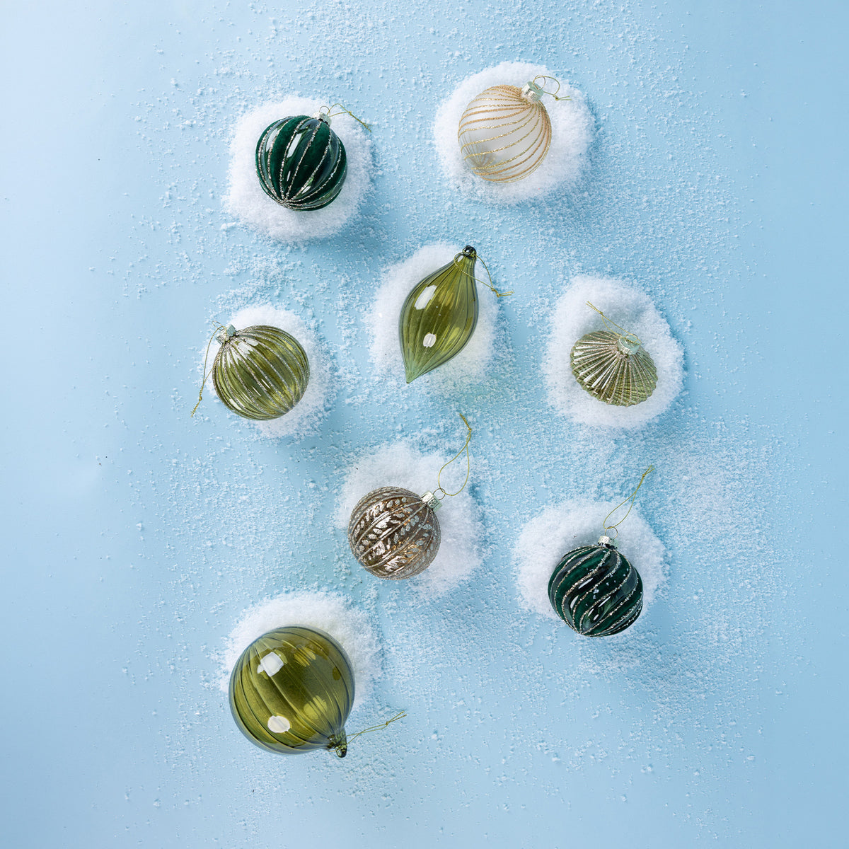 Assorted green, gold and champagne glass Christmas baubles featuring ribbed detailing, glitter accents and decorative shapes, arranged on a snowy blue background.