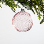 Pink round glass Christmas bauble featuring delicate swirling ribbed detailing, hanging from festive greenery against a white background.