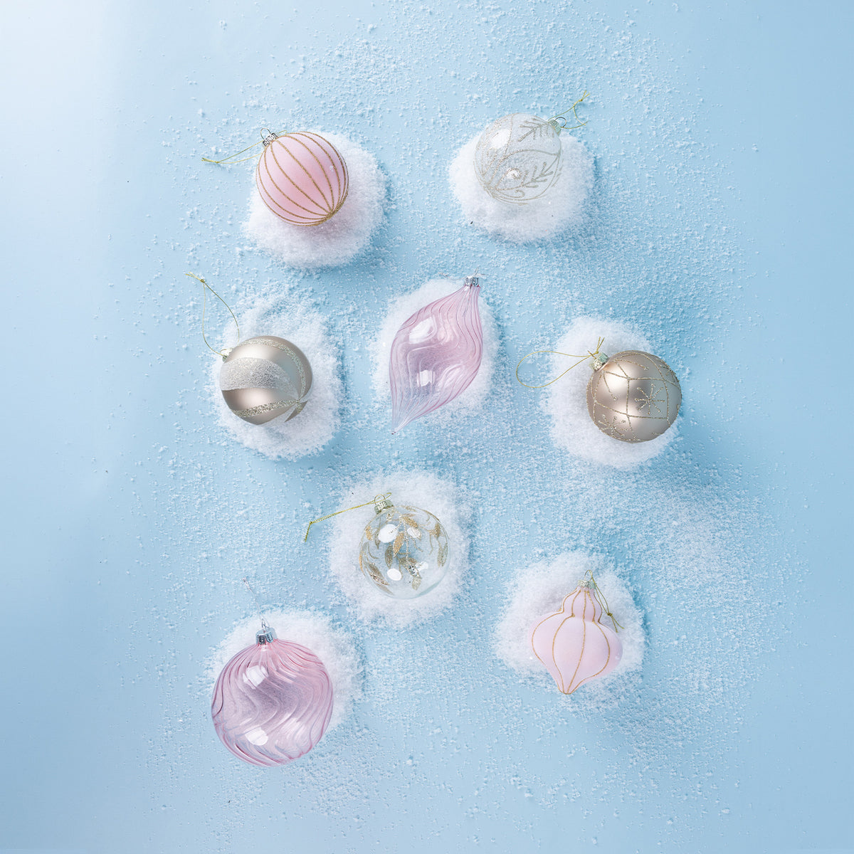 Assorted pink, white and champagne Christmas baubles featuring swirling glass designs, glitter accents and decorative patterns, arranged on a snowy blue background.