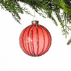 Round red glass Christmas bauble featuring vertical gold glitter stripes and a ribbed design, hanging from festive greenery against a white background.