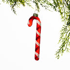 Red glass candy cane Christmas decoration featuring white glitter spiral stripes, hanging from festive greenery against a white background.
