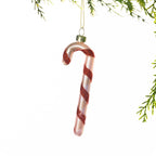 Pink glass candy cane Christmas decoration featuring red glitter spiral stripes, hanging from festive greenery against a white background.