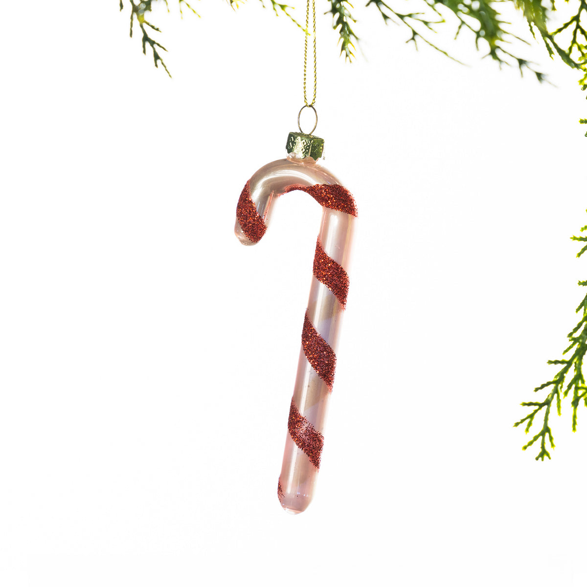 Pink glass candy cane Christmas decoration featuring red glitter spiral stripes, hanging from festive greenery against a white background.