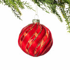 Red glass Christmas bauble featuring a swirling velvet design with gold glitter detailing, hanging from festive greenery against a white background.