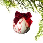 Light pink glass Christmas bauble featuring a red bow pattern and a large burgundy velvet bow, hanging from festive greenery against a white background.