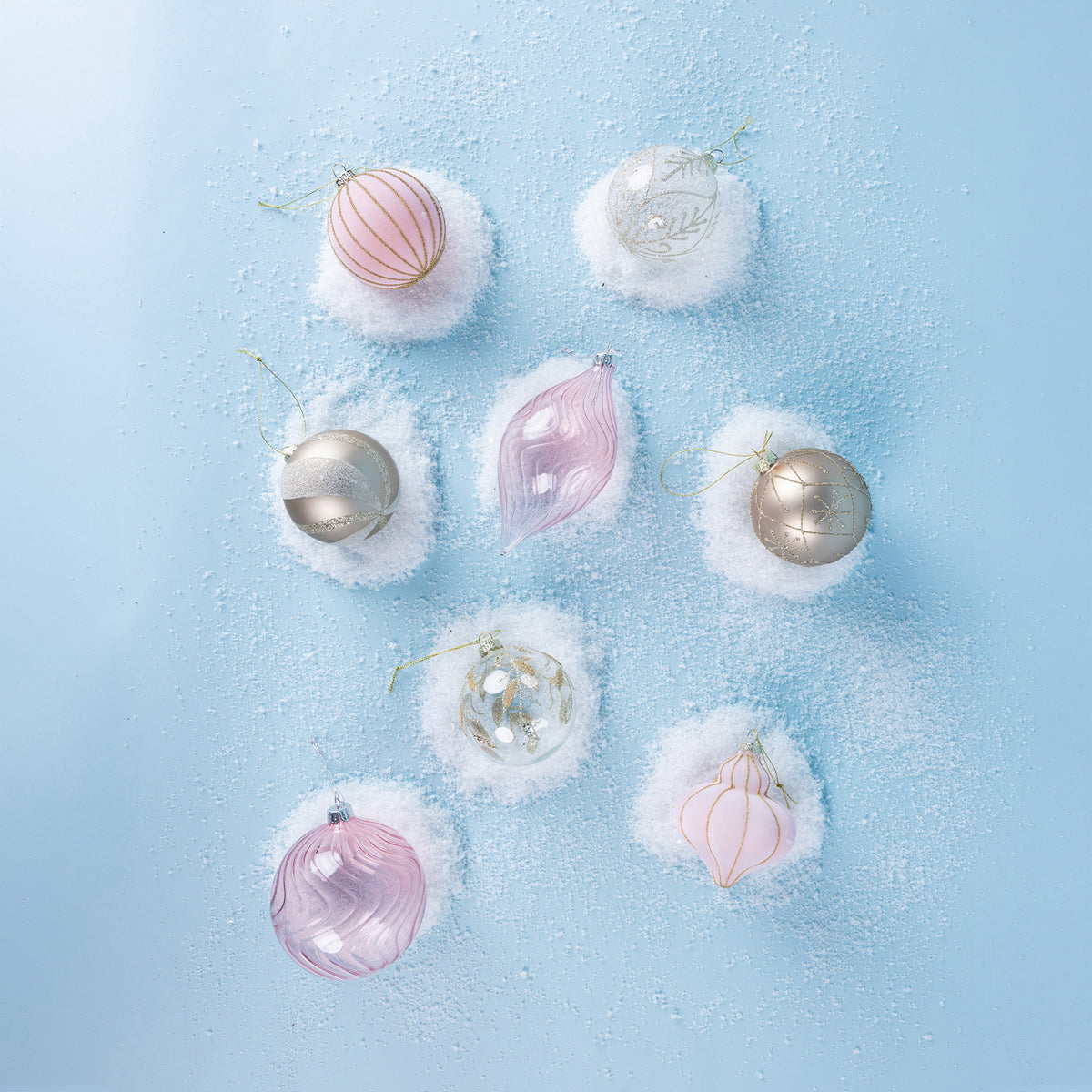 Assorted pink, silver and white Christmas baubles featuring glitter detailing and delicate patterns, arranged on faux snow against a light blue background.