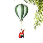 Green and white striped hot air balloon Christmas decoration with a miniature Santa holding a red sack suspended below, displayed against a white background with festive greenery.