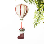 Cream and red striped hot air balloon Christmas decoration with a miniature stocking filled with presents suspended below, displayed against a white background with festive greenery.