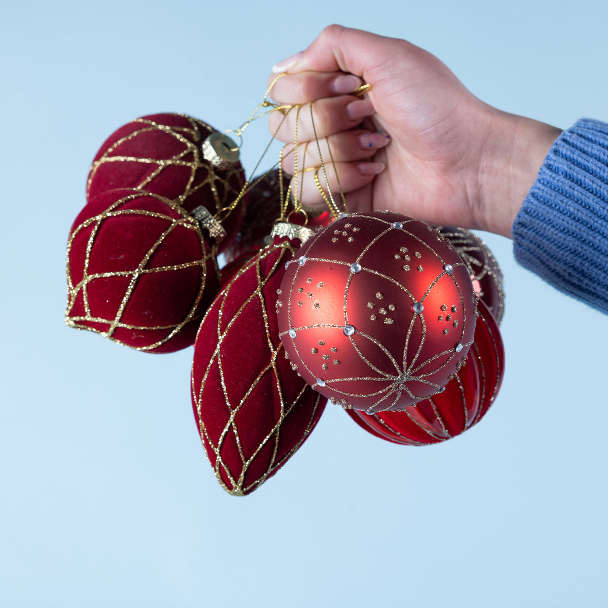 A hand holding a collection of red and burgundy Christmas baubles featuring velvet finishes, gold glitter patterns and decorative detailing against a light blue background.