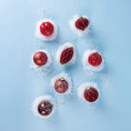 A collection of red and burgundy Christmas baubles with gold glitter detailing arranged on artificial snow against a light blue background.
