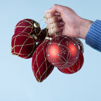 A hand holding a collection of red and burgundy Christmas baubles featuring velvet finishes, gold glitter patterns and decorative detailing against a light blue background.