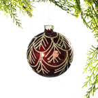 Deep red glass Christmas bauble featuring a gold glitter leaf pattern, hanging from festive greenery against a white background.
