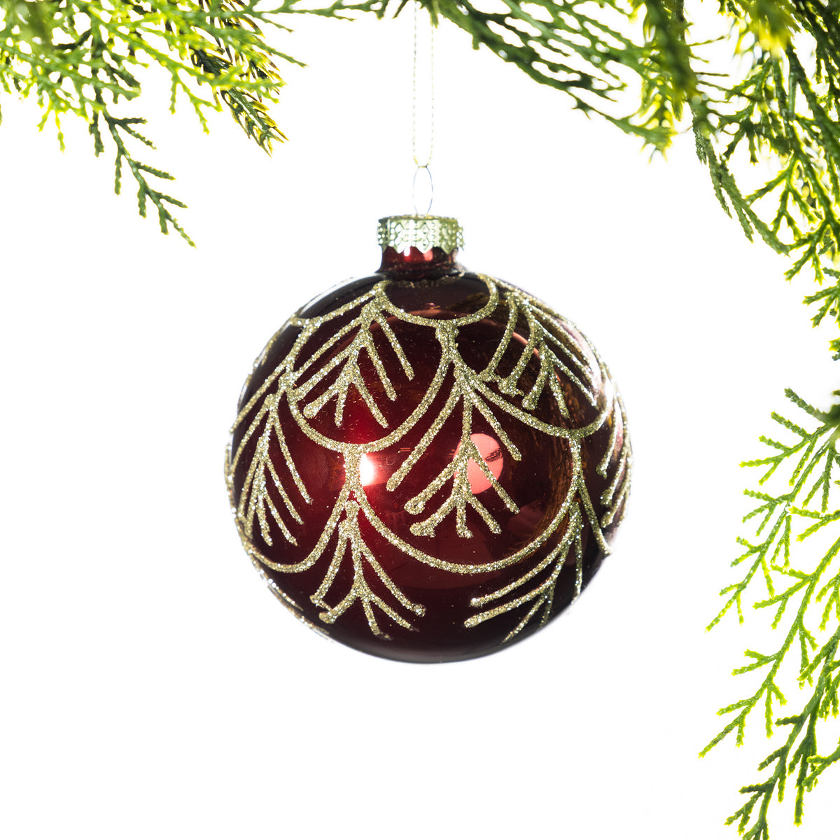 Deep red glass Christmas bauble featuring a gold glitter leaf pattern, hanging from festive greenery against a white background.