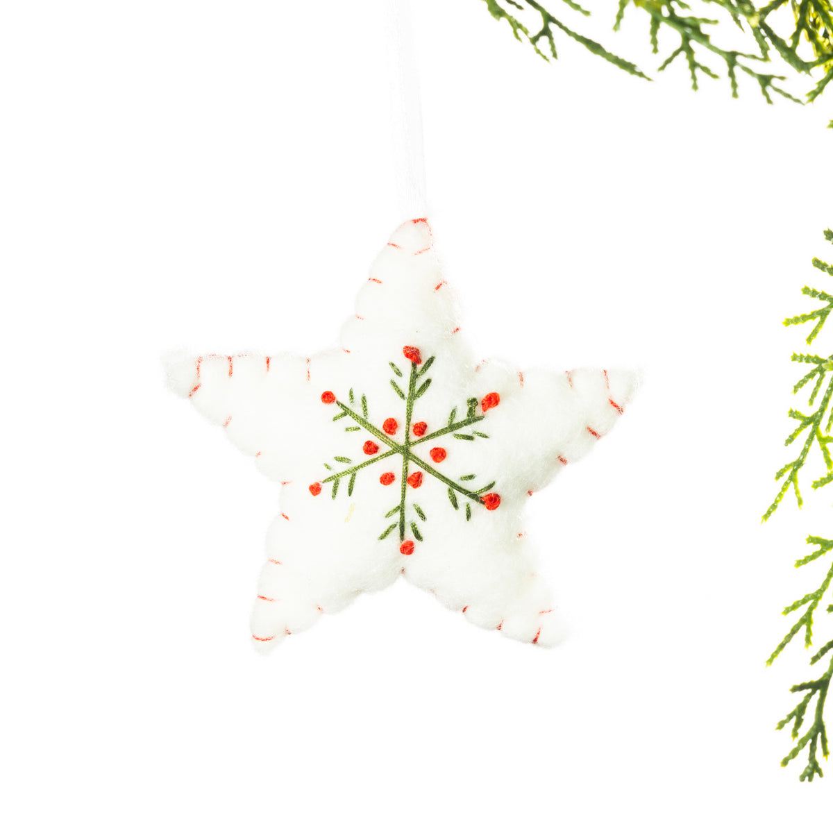 A white embroidered star hanging decoration featuring a green snowflake design with red accents and red stitching around the edges, suspended from festive greenery against a white background.