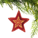 A red embroidered star hanging decoration featuring an intricate gold snowflake design and gold stitching around the edges, suspended from festive greenery against a white background.