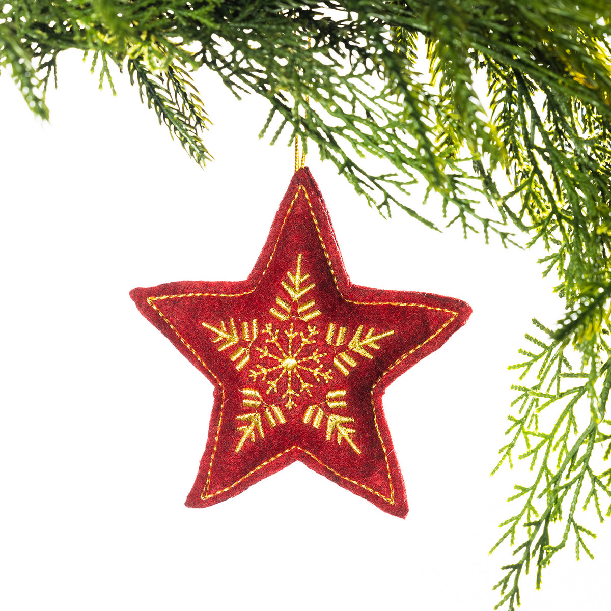 A red embroidered star hanging decoration featuring an intricate gold snowflake design and gold stitching around the edges, suspended from festive greenery against a white background.