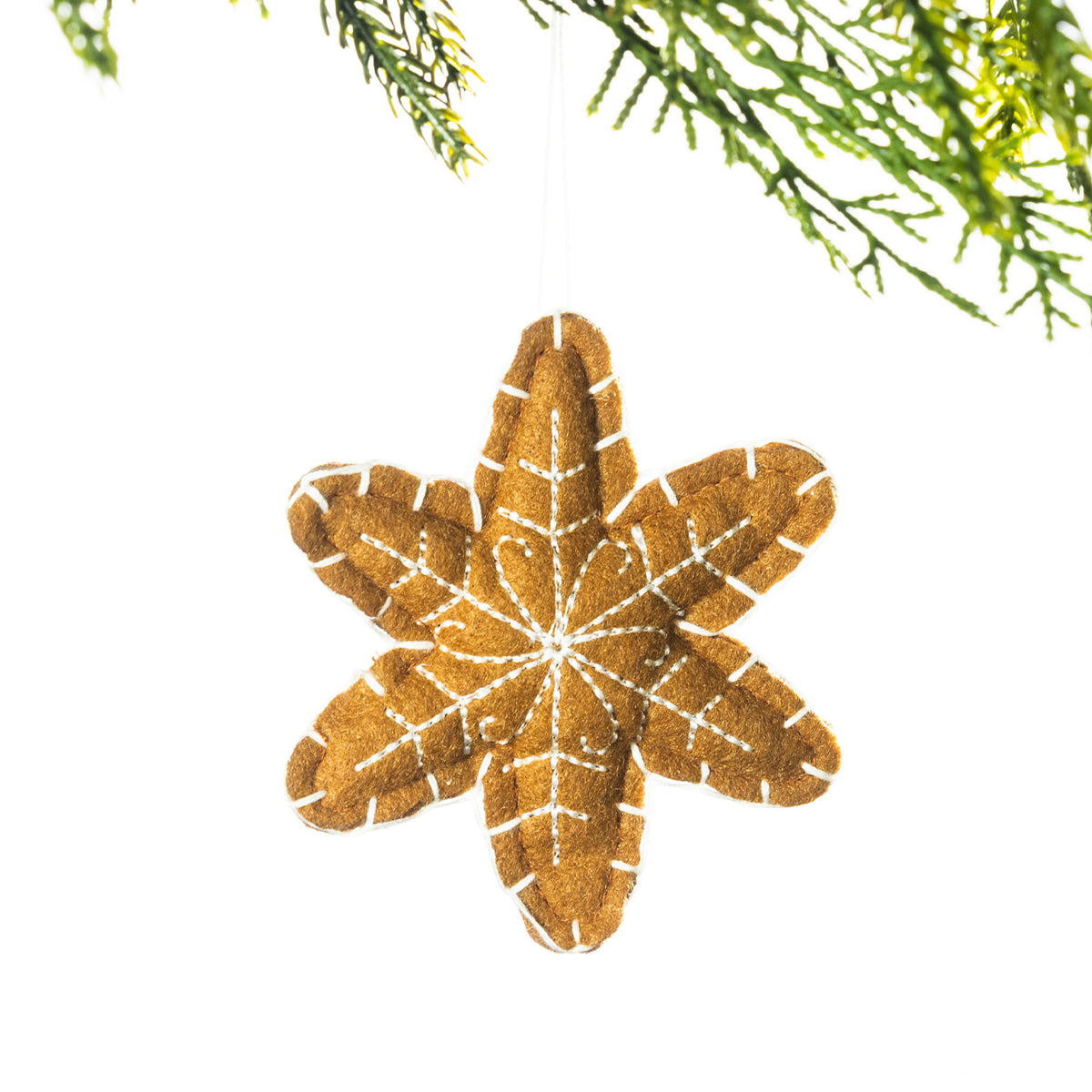 A gingerbread-coloured embroidered snowflake hanging decoration with white stitching, suspended from festive greenery against a white background.