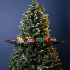 A decorated artificial Christmas tree with white snow-like dusting and warm yellow string lights, shown against a dark blue background. A miniature train set with Santa in a red sleigh and green cargo cars sits on a black shelf mounted behind the tree.