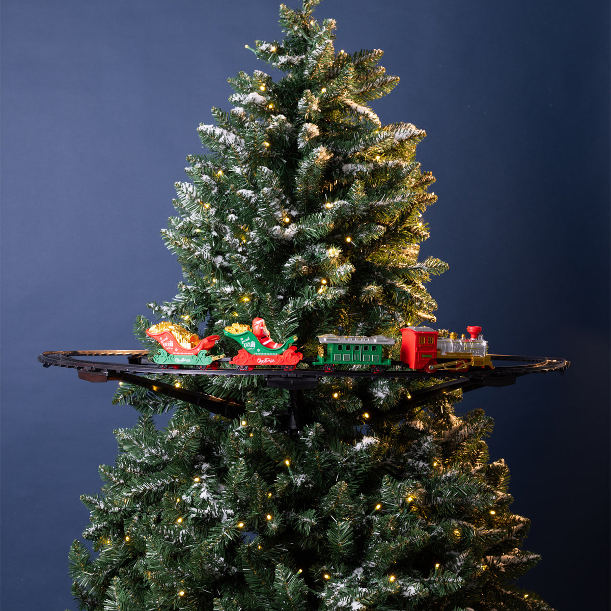 A decorated artificial Christmas tree with white snow-like dusting and warm yellow string lights, shown against a dark blue background. A miniature train set with Santa in a red sleigh and green cargo cars sits on a black shelf mounted behind the tree.