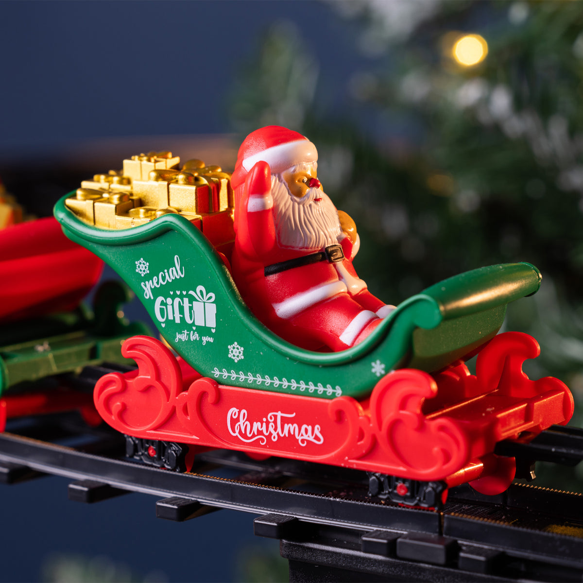 A festive Christmas Santa Claus sitting in a red and green sleigh loaded with gold gift boxes, photographed on toy train tracks with a blurred holiday background.