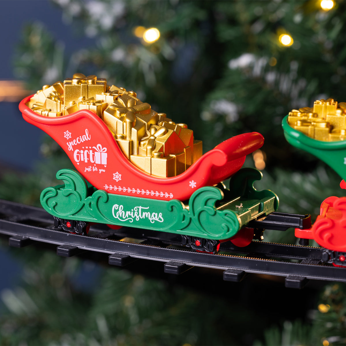 A festive Christmas-themed toy train set with a red and green sleigh carrying shiny gold gift boxes, running on black tracks against a blurred Christmas tree background.