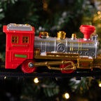 A festive toy locomotive train with a red cab, metallic silver boiler, and shiny gold details, running on a black track in front of a blurred Christmas tree background with warm lights.