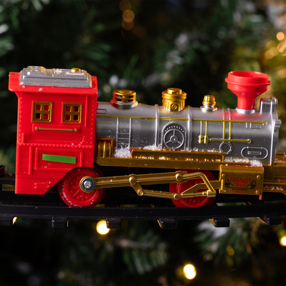 A festive toy locomotive train with a red cab, metallic silver boiler, and shiny gold details, running on a black track in front of a blurred Christmas tree background with warm lights.