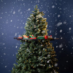 A decorated Christmas tree with snow-dusted branches and warm string lights, set against a dark blue snowy background. A small toy train set sits on a black shelf across the middle of the tree.