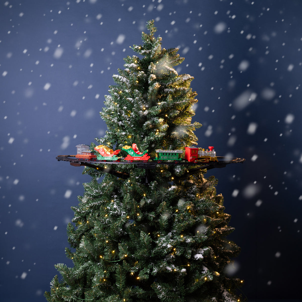 A decorated Christmas tree with snow-dusted branches and warm string lights, set against a dark blue snowy background. A small toy train set sits on a black shelf across the middle of the tree.