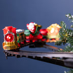 A festive Christmas toy train set on a curved black track, featuring a red and gold locomotive pulling holiday-themed cars, with a Santa figure and a glowing sack of coins in the background. A pine branch with warm lights frames the right side against a dark blue backdrop.