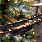 Close-up of a black plastic train track section, photographed against a blurred Christmas tree with warm lights.