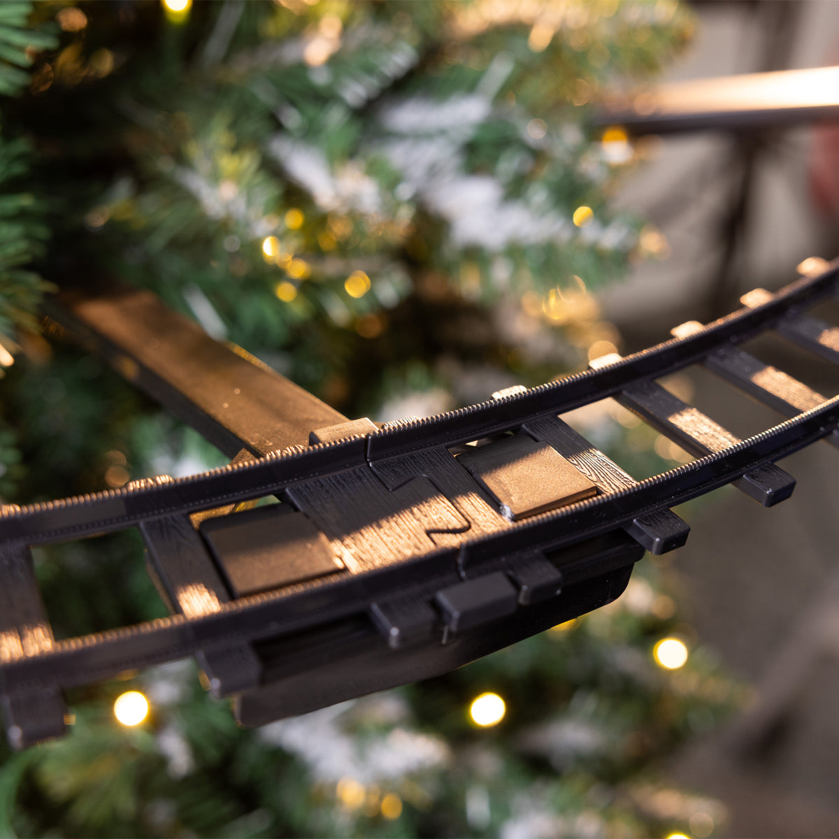 Close-up of a black plastic train track section, photographed against a blurred Christmas tree with warm lights.