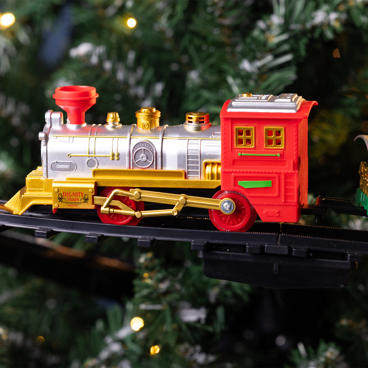 A toy model locomotive train with a red caboose, silver boiler, and gold details, displayed on a black track against a blurred Christmas tree background with warm lights.