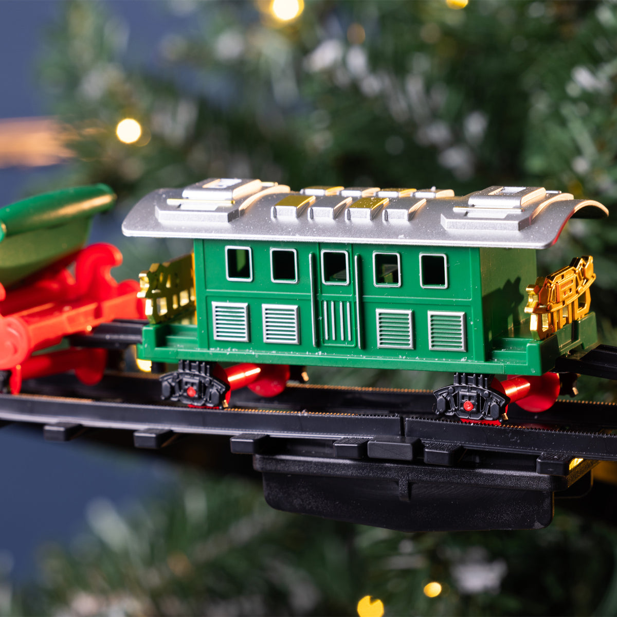 A green toy train passenger car with red and gold details, mounted on a black track, photographed against a blurred Christmas tree background with warm lights.