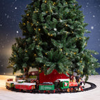 A festive Christmas tree scene with a Christmas train track circling the base of a decorated artificial Christmas tree. Under the tree are wrapped presents, a red gift box, and small holiday figures, set against a snowy night backdrop with falling snow.