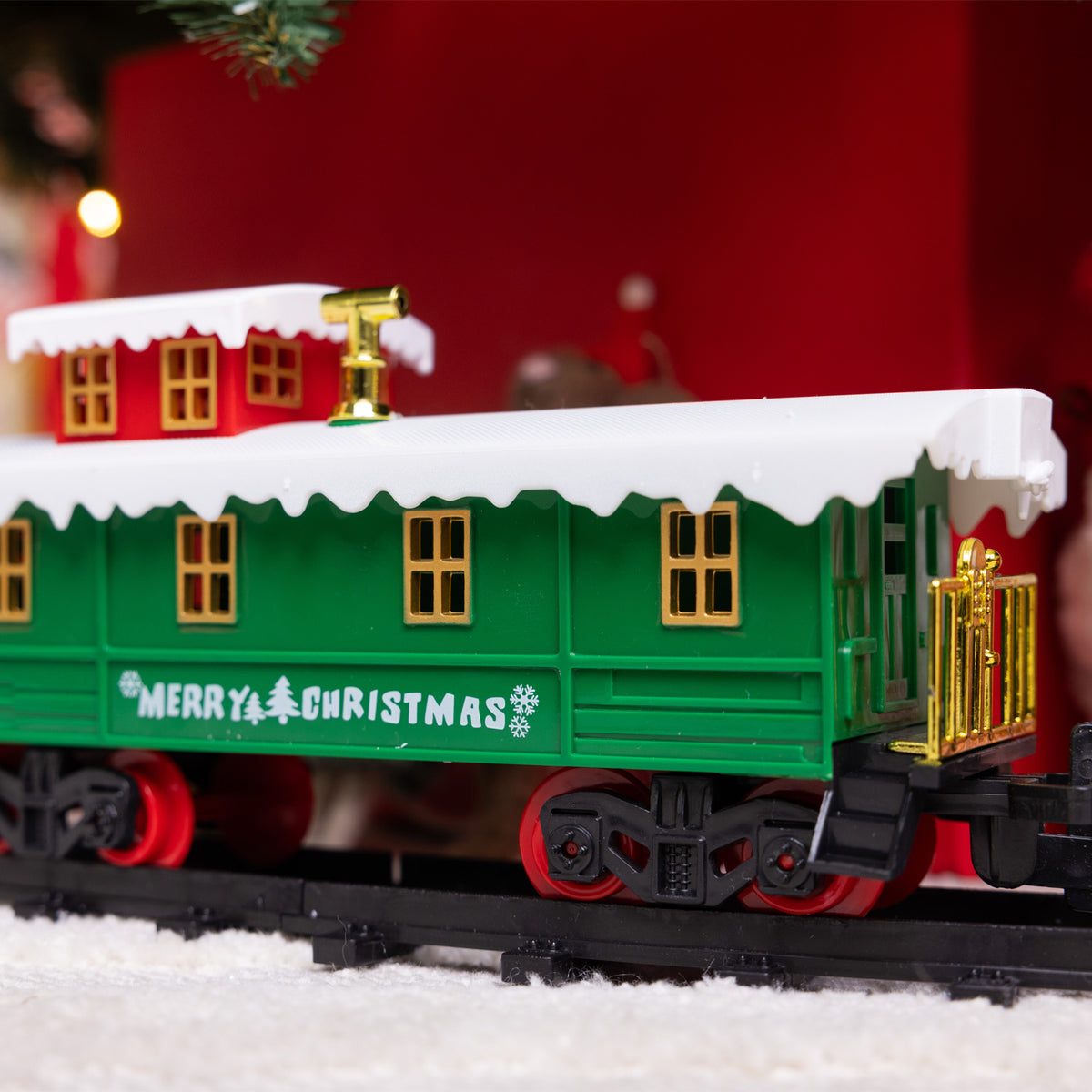 A green Christmas-themed train car with 'Merry Christmas' written on the side, featuring white snow-dusted roofs, gold accents, and red wheels, photographed on a snowy surface with a blurred festive background.