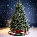 A decorated artificial Christmas tree with snow-dusted branches, warm yellow string lights, and a red base, surrounded by a small train track with a green and red locomotive. Presents sit at the base, and a snowy scene is set against a dark blue background with falling snow.