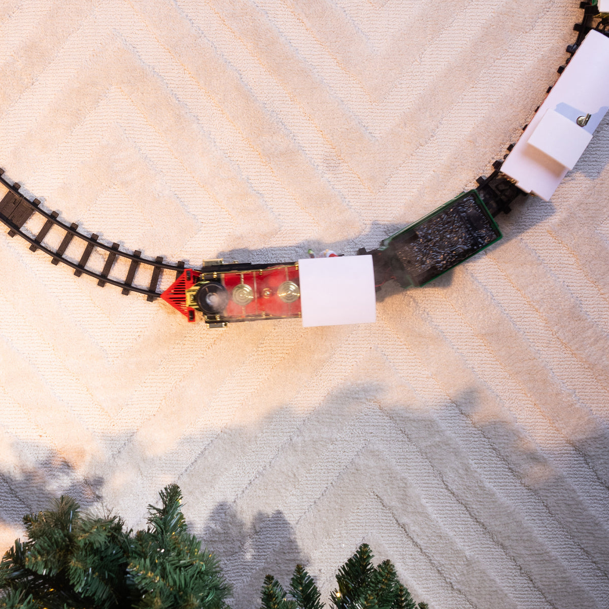 A model steam train set with a red locomotive and black cargo cars on curved tracks, photographed from above on a textured beige surface with small green trees at the bottom.
