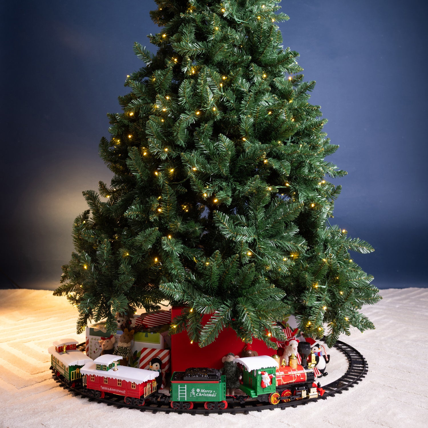 A decorated artificial Christmas tree with warm white string lights, surrounded by a small Christmas train on a circular track and wrapped gifts at its base, set against a dark blue gradient background.