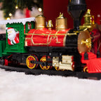 A festive Christmas electric train locomotive with a red body, gold accents, a green cab, and a small Santa Claus figure in the window, shown on a snowy surface with a blurred holiday background.