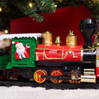 A festive Christmas-themed decorative steam locomotive train with a bright red body, gold brass details, a green cab, black wheels, and a Santa Claus figure waving from the window. The train is set on a snowy white surface under a decorated Christmas tree with warm lights.