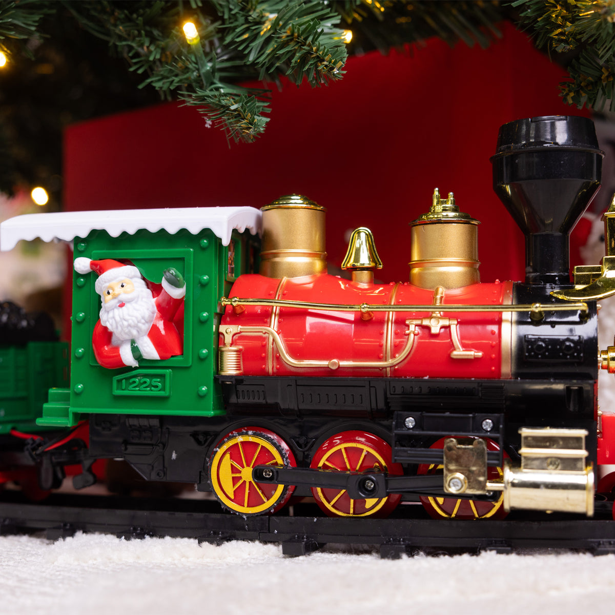 A festive Christmas-themed decorative steam locomotive train with a bright red body, gold brass details, a green cab, black wheels, and a Santa Claus figure waving from the window. The train is set on a snowy white surface under a decorated Christmas tree with warm lights.