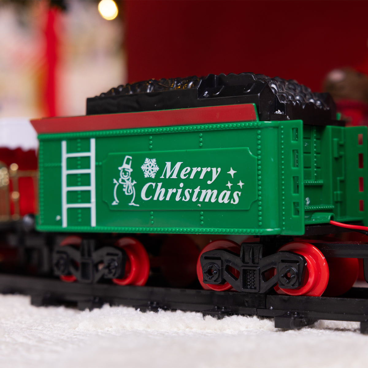 A green train car on a black track, decorated with a white Christmas snowman graphic and the words 'Merry Christmas' in white. The car is filled with black cargo and has red wheels, set against a blurred festive background with red and warm lights.